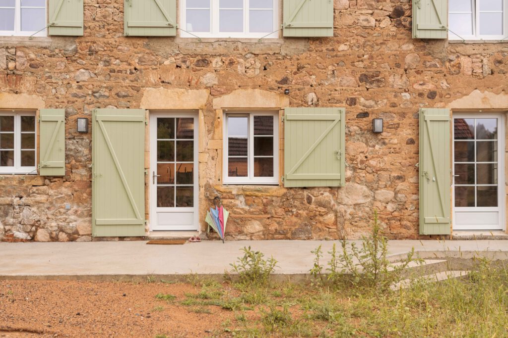 Photographie de la façade d'une maison en rénovation dans le Beaujolais. Elle est en pierre avec des volats vert sauge.