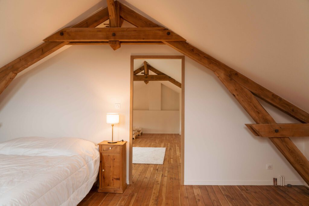 Photographie d'une enfilade de chambres en soupente suite à la rénovation d'une maison d'habitation dans le Beaujolais. Les murs sont blancs et les poutres en bois apparent. L'atmosphère est douce.